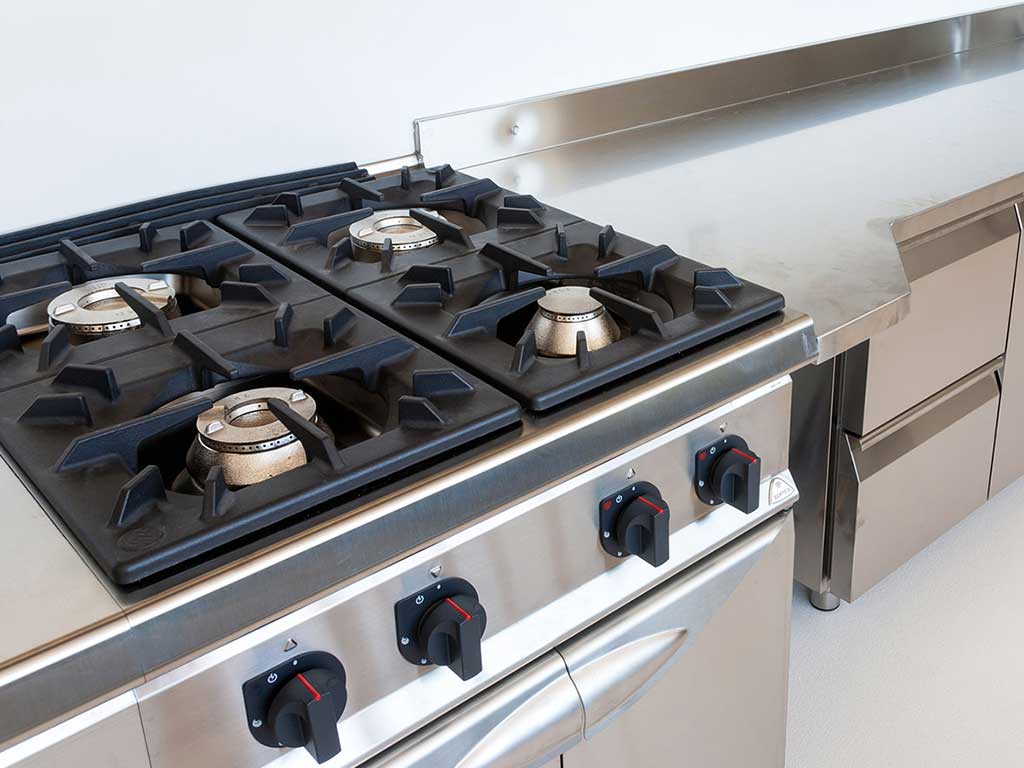 Eine Nahaufnahme eines sauberen, vierflammigen Gastronomie-Gasherds aus gebürstetem Edelstahl. Die massiven schwarzen gusseisernen Topfträger sind deutlich erkennbar, ebenso wie die robusten schwarzen Drehschalter mit roten Markierungen an der Vorderseite des Geräts.